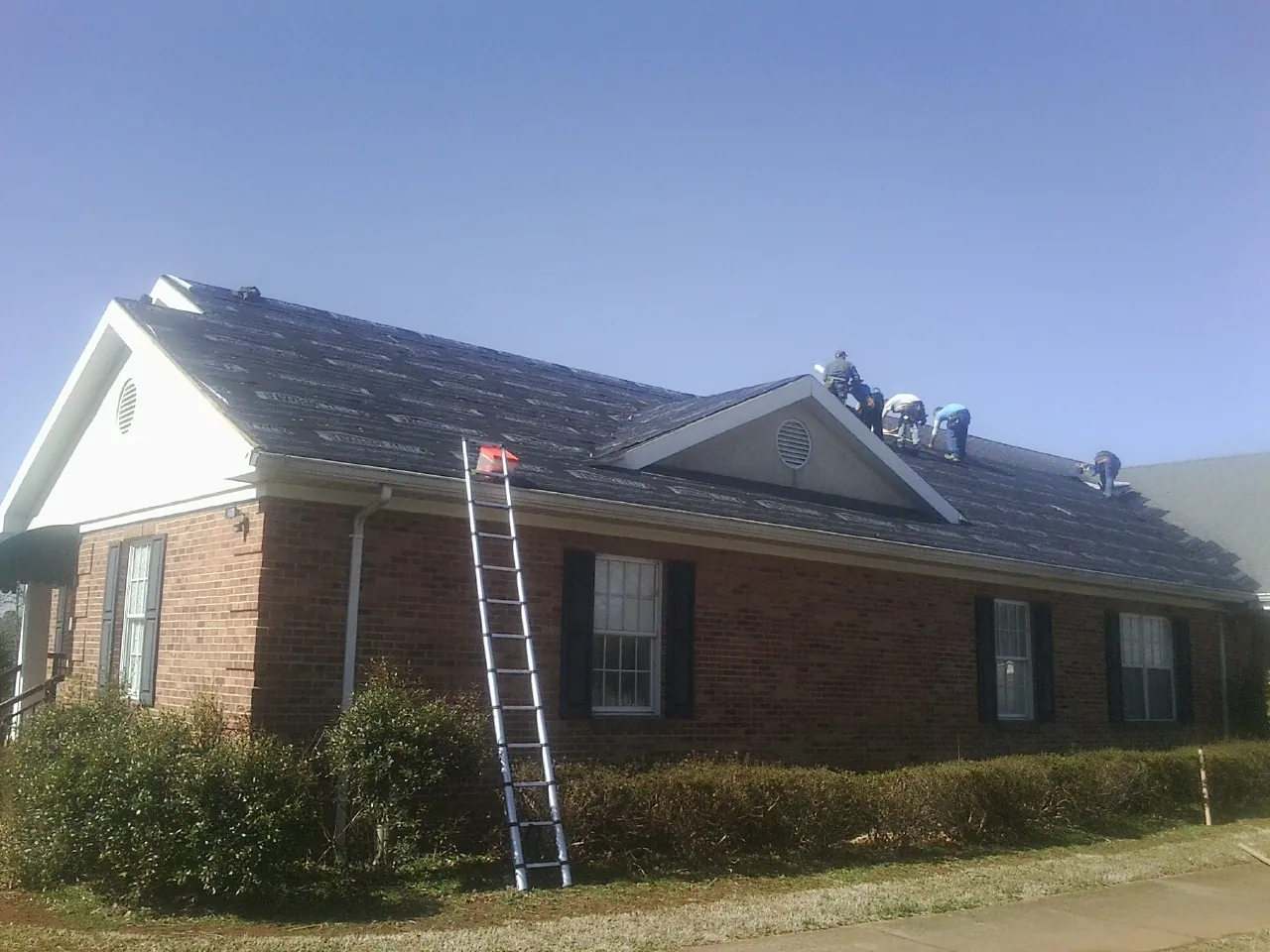 Crew tearing shingles off a brick ranch roof with a ladder set — Emergency Roof Repair work by our crew in Clay