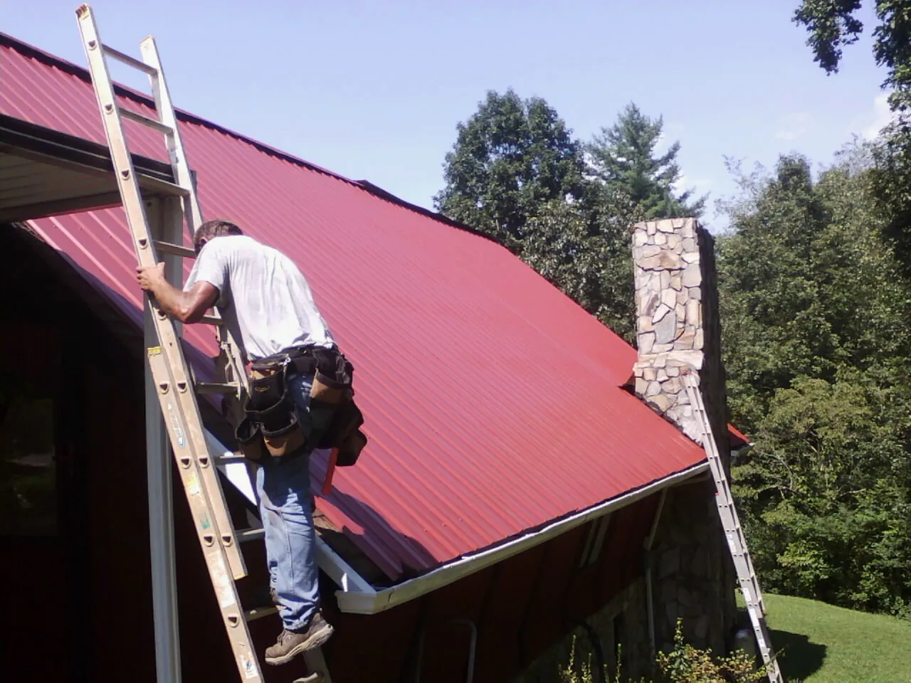 Roofer on a ladder beside a red metal roof and stone chimney — Soffit & Fascia Repair by Roofing Brothers in Clay
